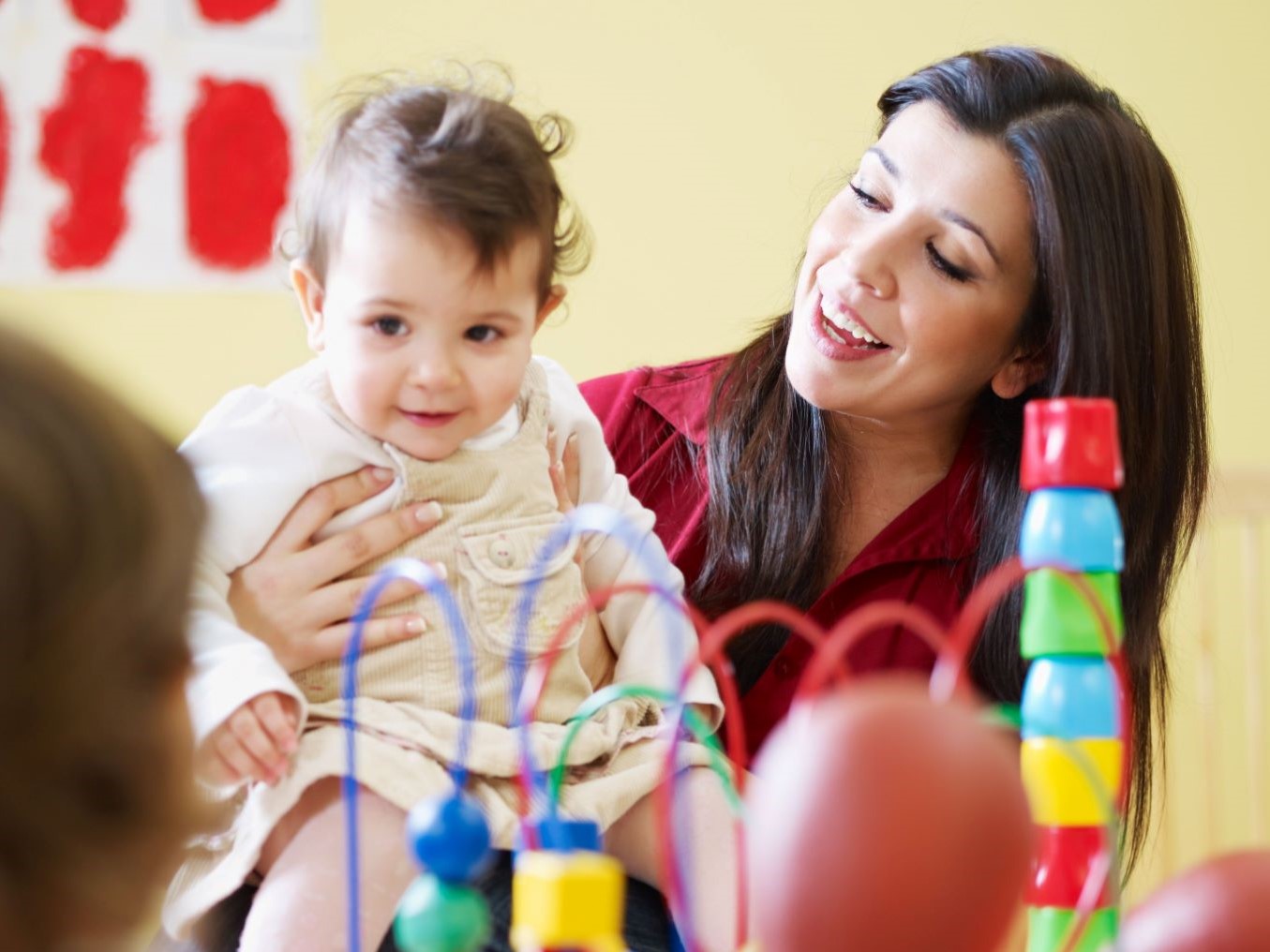 Femme souriante tenant une petite fille sur ses genoux. Au premier plan, des jeux d'éveil colorés