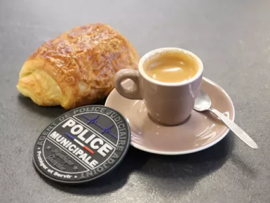Une tasse de café, un pain au chocolat et un écusson de la Police municipale posés sur une table