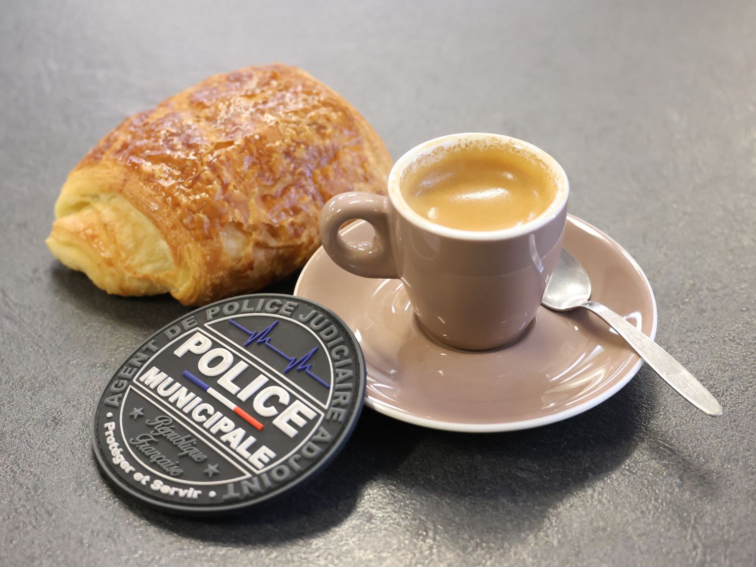 Une tasse de café, un pain au chocolat et un écusson de la Police municipale posés sur une table